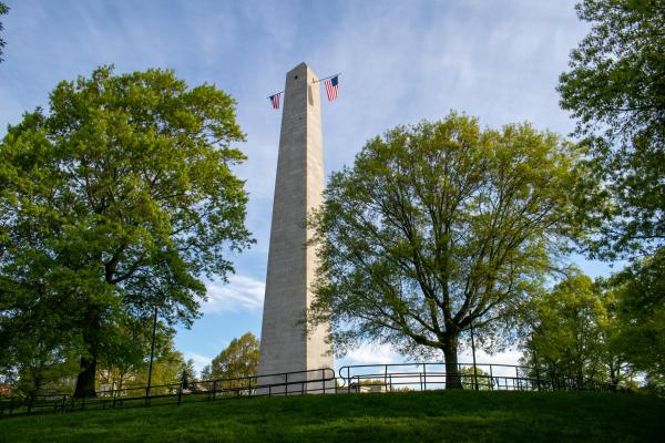 A tall white obelisk stands amongst leafy trees. Two colonial American flags hang from its windows