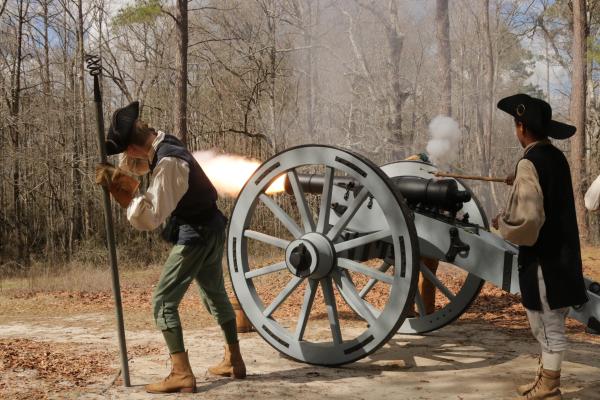 Four people in period clothing standing next to a cannon with a fireball coming out of the barrel