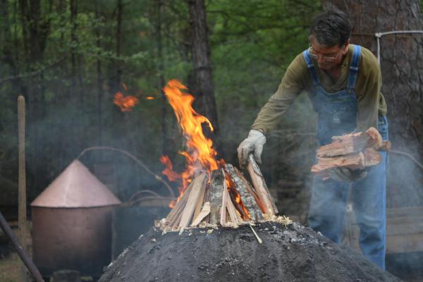 Man loading wood on to a dirt mound with a fire burning.