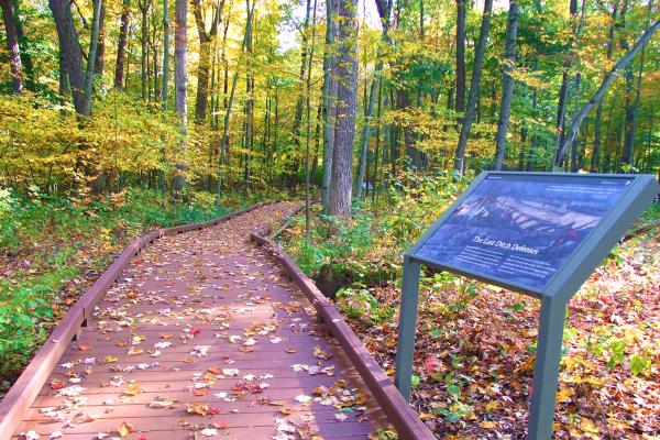 An informational sign sits beside a wooden walkway winding amid early autumn trees.