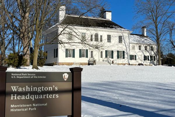 A white colonial building with snow in the foreground and a blue sky and trees in the background