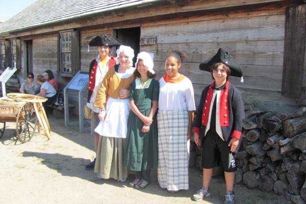 Five children stand in a row smiling. They all have different 18th Century accessories on.