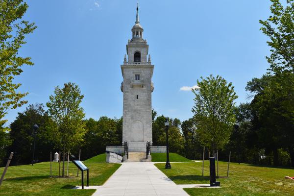A white marble steeple-looking tower stands amongst trees in a community park.