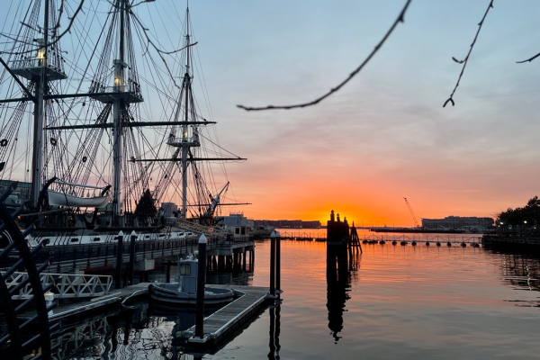 A three-masted ship sits in still water as the sun sets over Boston Harbor.