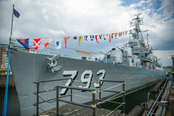 A gray naval destroyer sits next to a dock. It has "793" on the side and colorful signal flags.
