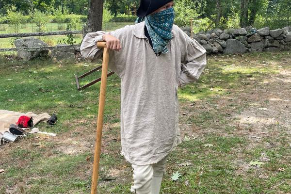 A colonial man with a cloth over his face stands holding a shovel