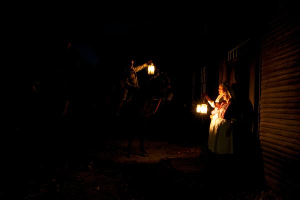 Two women in colonial clothing stand with a lantern in the darkness near a man on a horse.