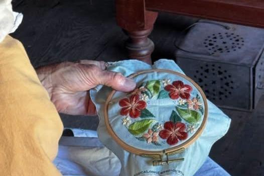 A volunteer in colonial dress practices cross stich. She is stitching a flower design.