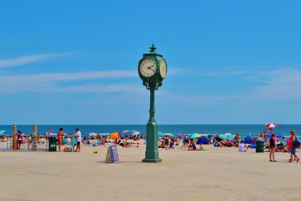 Green clock on boardwalk of ocean beach park site with people, umbrellas and blankets on sand