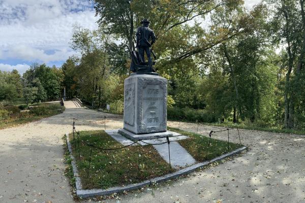 The state of the Minute man stands atop a stone base. The wooden North Bridge is seen in the distanc