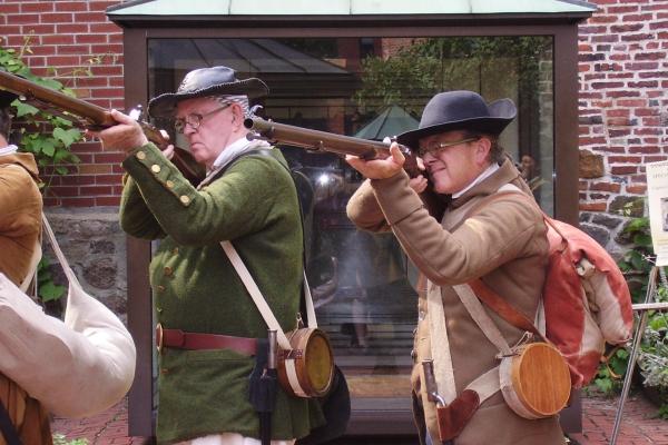 two men in colonial militia clothing pointing muskets at the camera