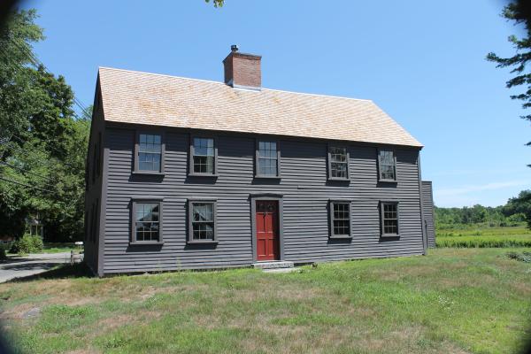 A wooden two story colonial house with dark paint sits in a field of green grass
