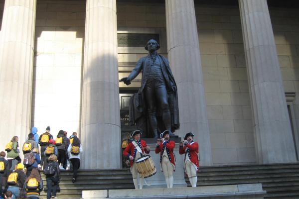 Three people in reenactment clothing hold instruments in front of a stone building and giant statue.