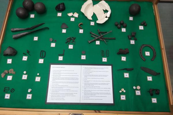 A collection of artifacts such as musket balls, nails, a horseshoe, a broken plate and cannon balls.