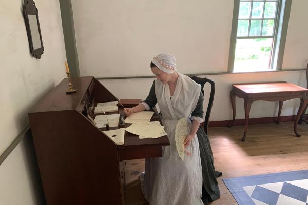 A woman in colonial clothing sits at a desk writing