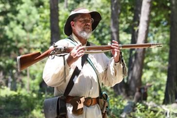 A living historian holds a weapon with trees behind him.