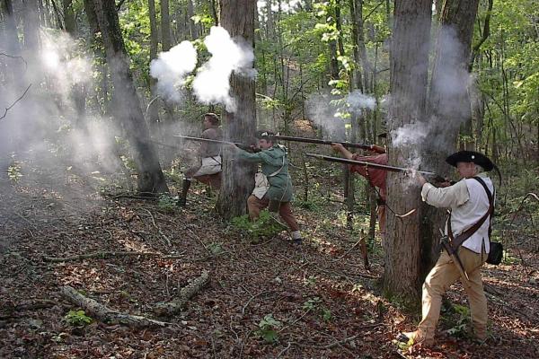 Living historians fire muskets with gray smoke coming from them. They are in the woods.