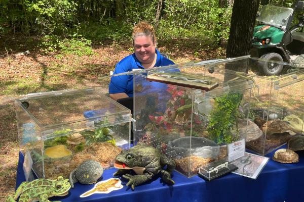 Woman in a blue polo shirt sits at a table with a variety of amphibians and reptiles in glass cages