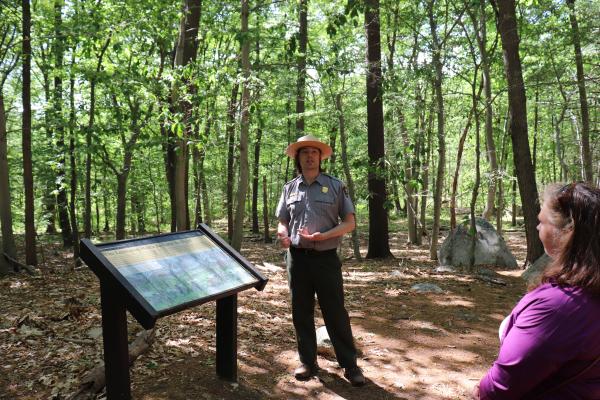 Man in green and gray NPS uniform standing next to an exhibit panel talking with visitors