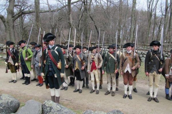 Several Colonial Revolutionary War militiamen stand in a line with muskets.