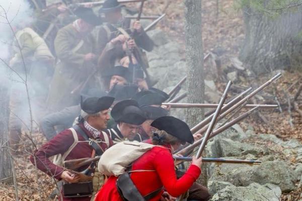 Revolutionary War Colonial soldiers take cover behind a low stone wall and fire their muskets