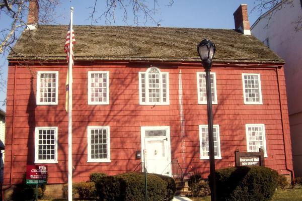 A red historic house with white trim and twin brick chimneys stands behind a flagpole and walkway lined with bushes.