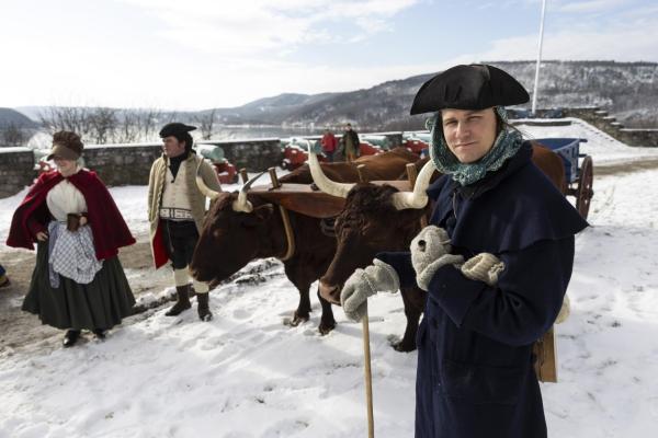 A man in 18th century clothing stand holding a pipe next to two oxen in winter time.