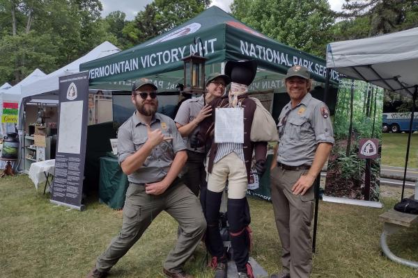 Three people in gray shirts and green pants post together with a mannequin.