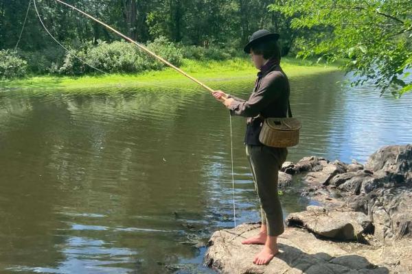 An 18th century man stands at a river fishing. He has a wicker basket on his hip and no shoes on.