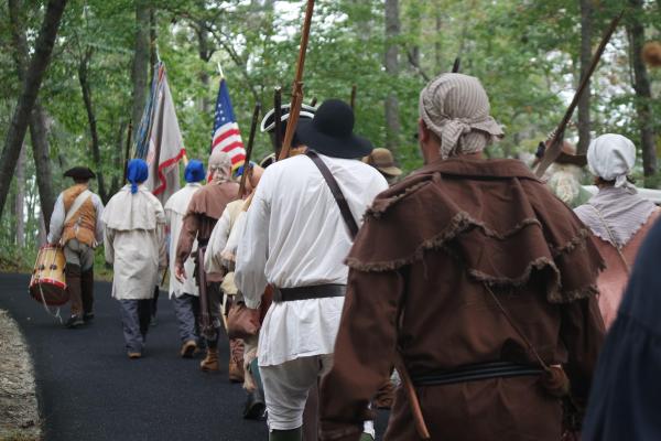 People in historical costumes march behind an American flag and a drummer.