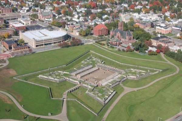 A view of a star-shaped wooden fort in a field of verdant green, from above. A city surrounds it.