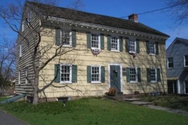 A yellow colonial‑style house with green shutters and a steep dark roof stands behind a leafless tree, with a stone walkway leading to its green front door.