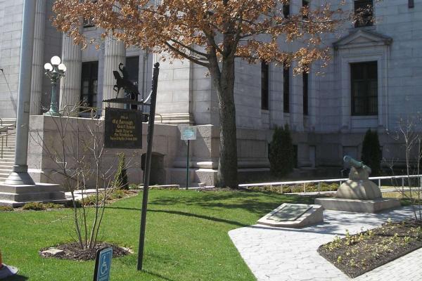 A light‑stone courthouse stands behind a grassy area with a tree, a small cannon monument, and a historical marker noting the site of an early borough courthouse built before the Revolution.
