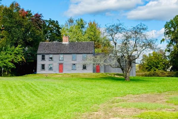 A wooden colonial era salt box house with red painted door sits in a green field of grass