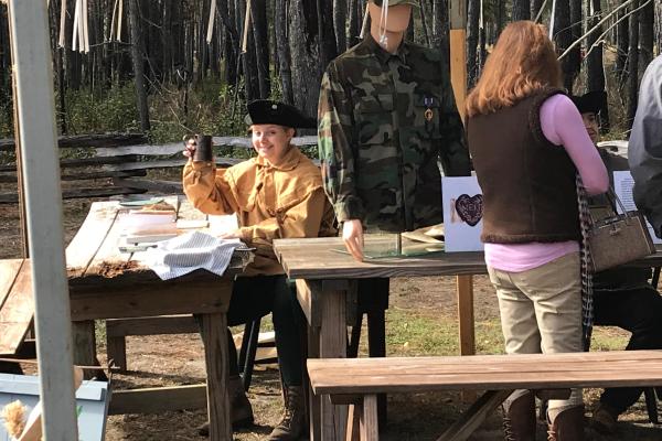 Person wearing colonial clothing sitting at a wooden table with a mannequin on the table.