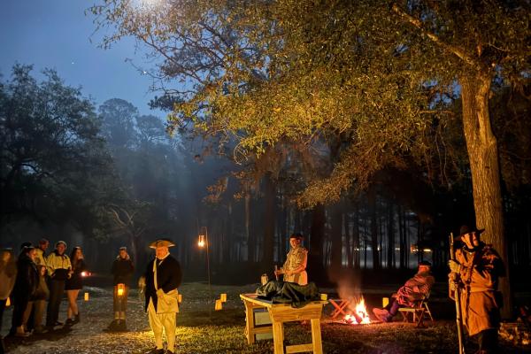 People dressed in colonial clothing outside at night with lanterns and a full moon illuminating them