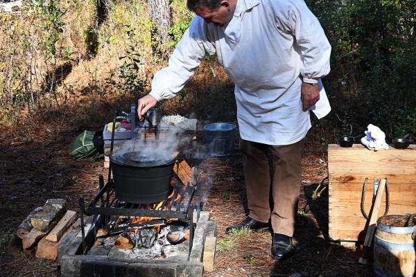 Man in a white smock bending over a campfire dipping a candle