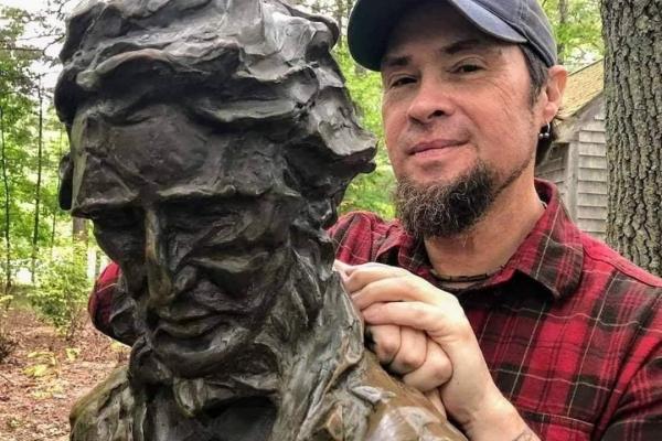 A man in a plaid shirt leans on a bronze monument of Henry David Thoreau