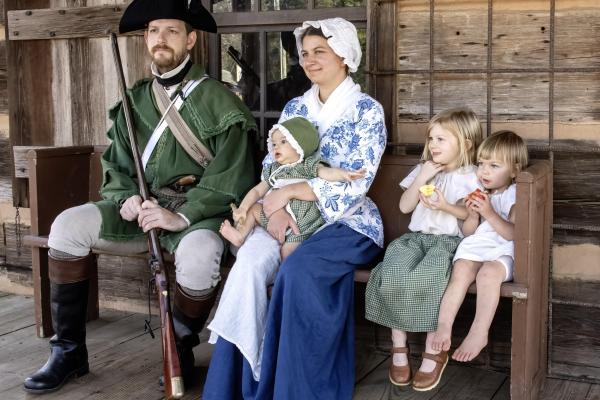 A family of 5 wearing 18th century clothing are sitting on a bench by a log cabin.
