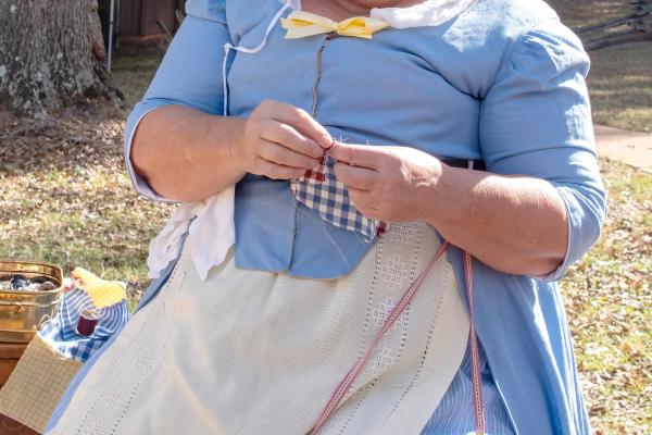 A woman in 18th century clothing is knitting and smiling.
