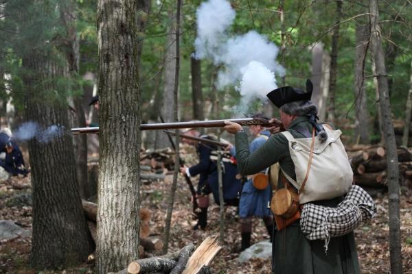 A militia soldier leans against tree and fires a musket