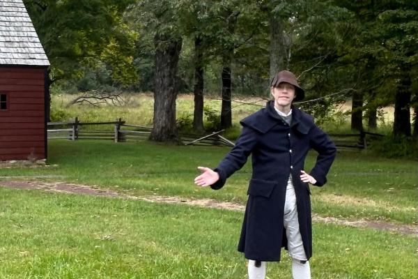 A man in 18th century period clothing stands in front of a campfire pit.
