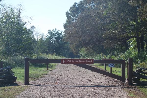 A brown metal gate crosses a gravel road, flanked with wooden fence and tall grass