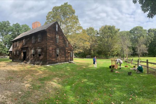 A historic wooden house in a field with people dressed in colonial clothing outside, and a cider pre