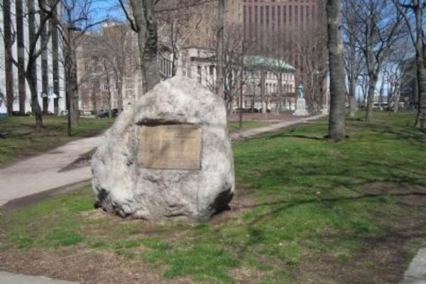 A large boulder with a metal plaque stands in a grassy urban park surrounded by trees and tall buildings.