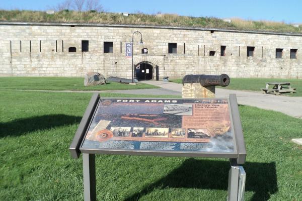 A stone fort wall with an arched entrance rises behind a grassy area where an informational sign, an old cannon, and a picnic table sit under a bright blue sky.