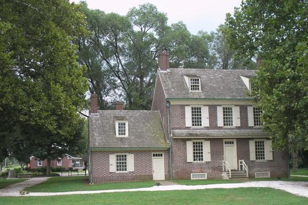 A brick colonial‑style house with cream shutters and multiple chimneys stands amid green grass and tall trees.