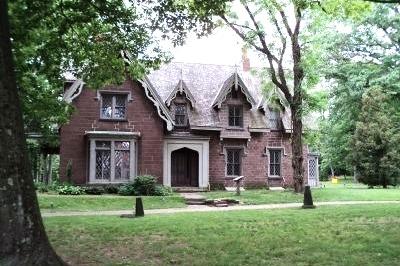 A large brick Gothic‑style house with steep gables and tall narrow windows stands among mature trees on a grassy lawn.