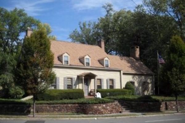 A tan colonial‑style house with dormer windows, brick chimneys, and a columned porch stands behind trimmed hedges and a stone retaining wall, with an American flag flying beside it.