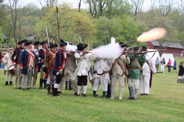 A line of Revolutionary War re-enactors in period uniforms fire muskets during a demonstration at Jockey Hollow. Smoke and muzzle flashes rise as the volley is discharged. Behind them, more re-enactors stand at attention with fixed bayonets.